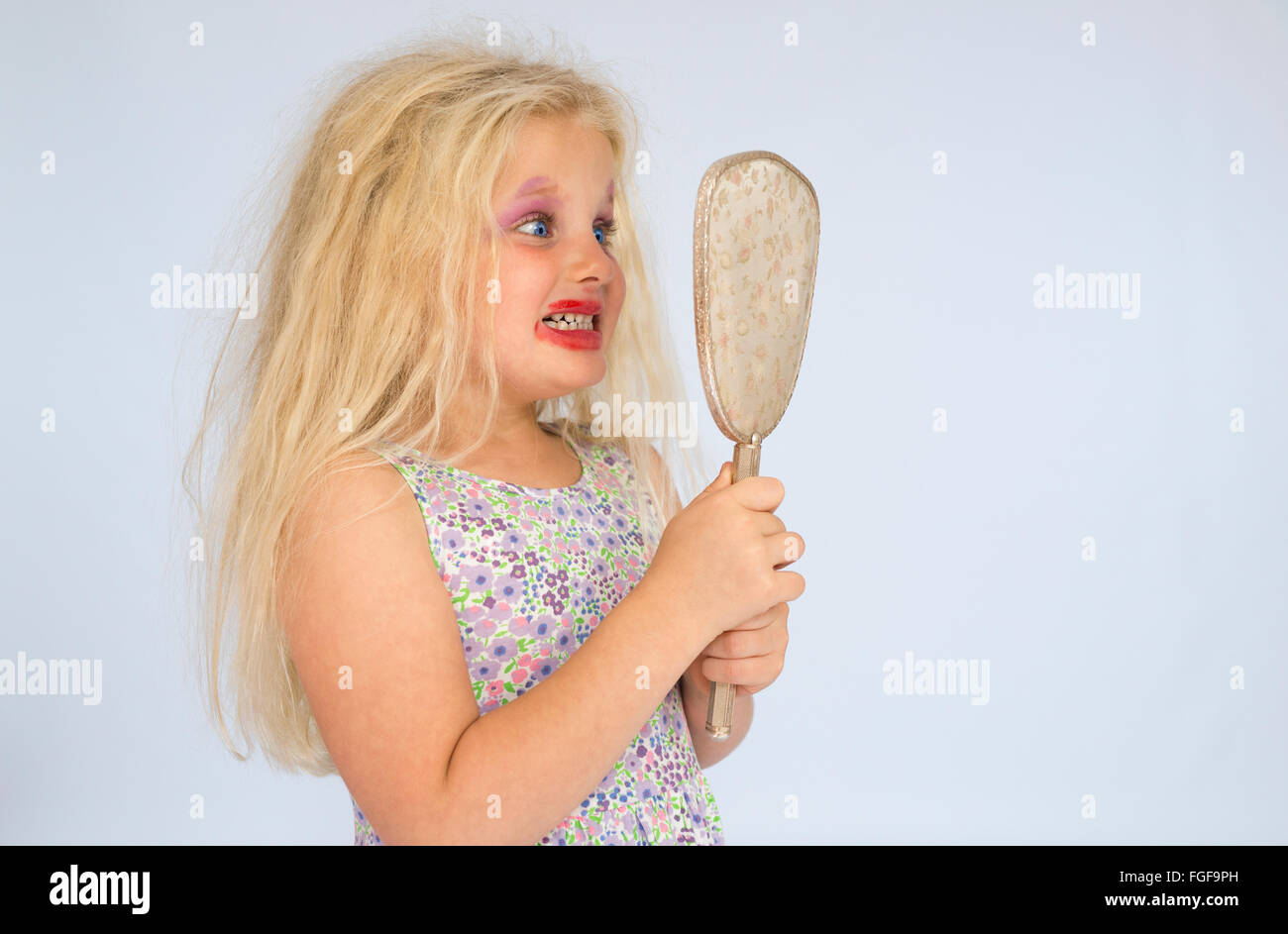 Muchacha con cabello rubio vistiendo maquillaje manchado mirando sorprendido a sí misma en el espejo Foto de stock