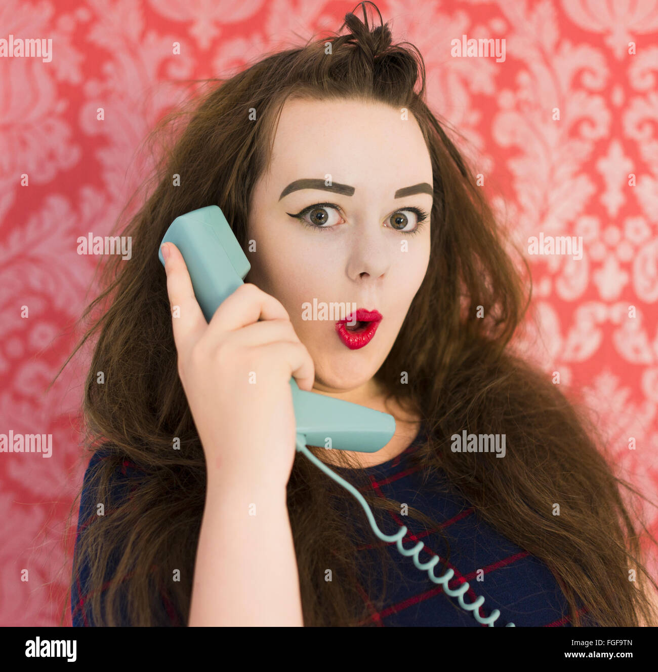 Adolescente sujetando un teléfono azul retro mirando sorprendido contra un fondo de papel tapiz vintage rojo Foto de stock