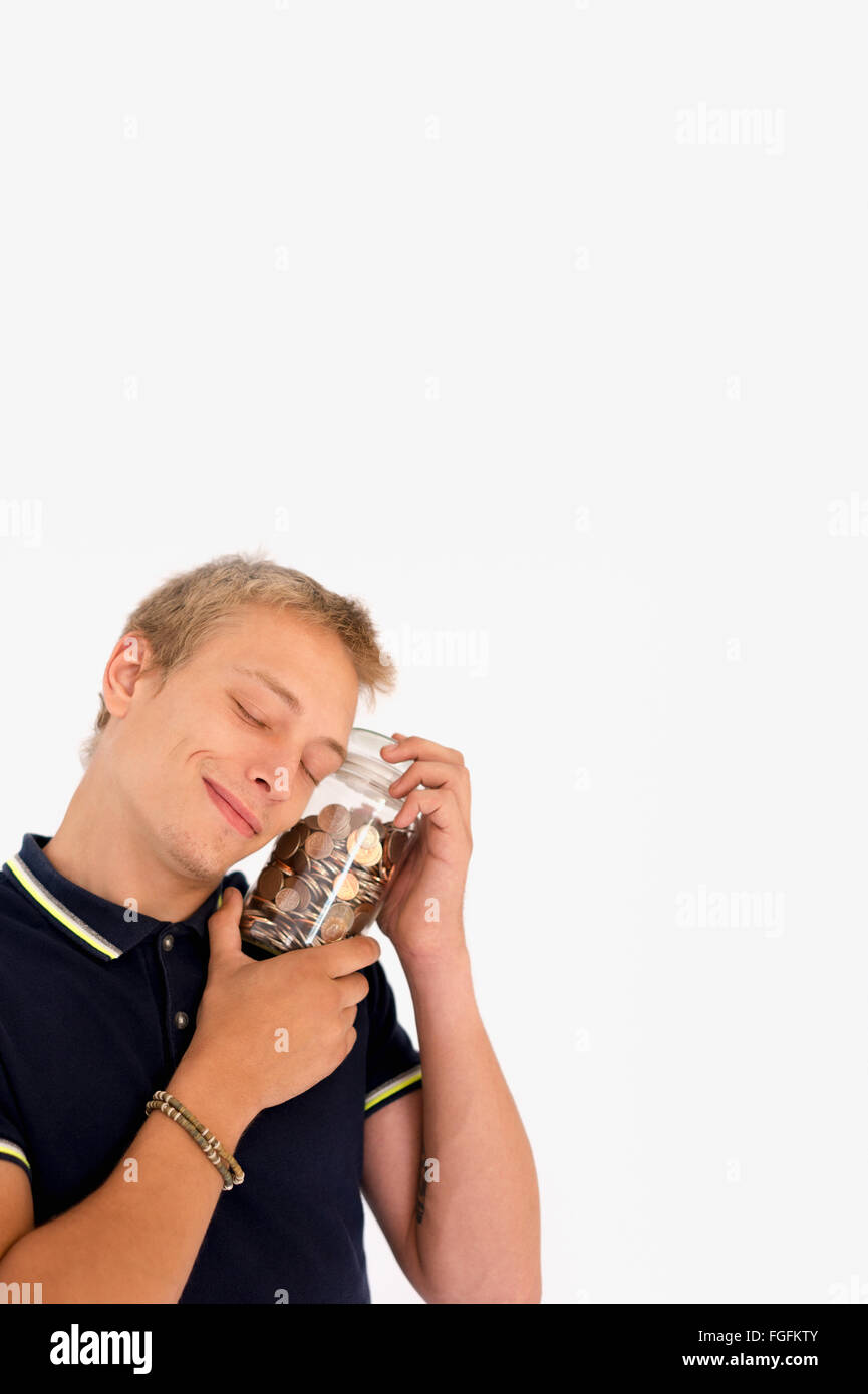 Hombre sujetando un tarro de monedas británicas mixtos cerca de su mejilla, sonriendo Foto de stock