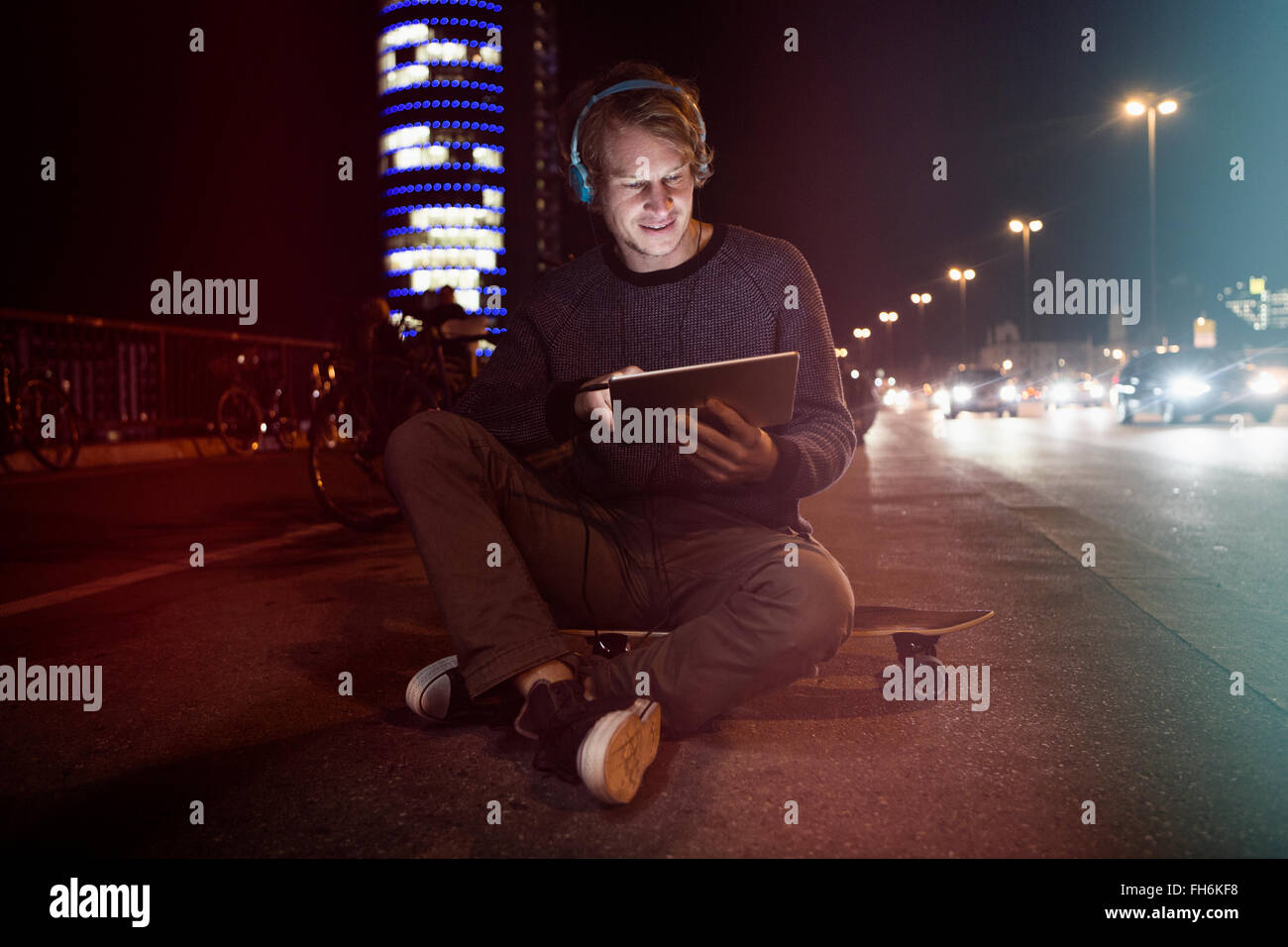 Alemania, Munich, Hombre con auriculares sentado en su patineta usando tableta digital por la noche Foto de stock