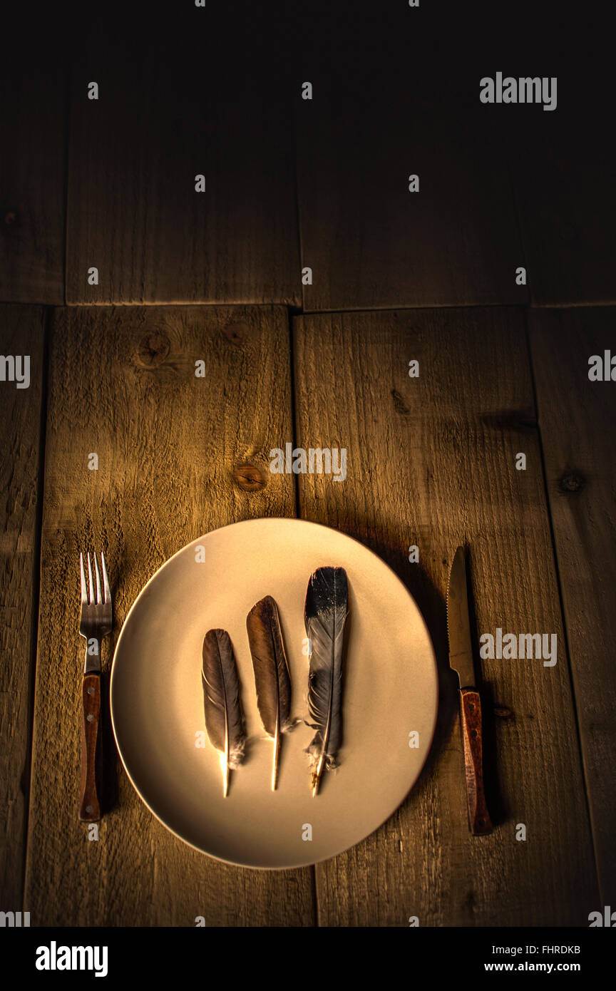 Bodegón con plumas en un plato y tenedor y cuchillo sobre mesa de madera Foto de stock