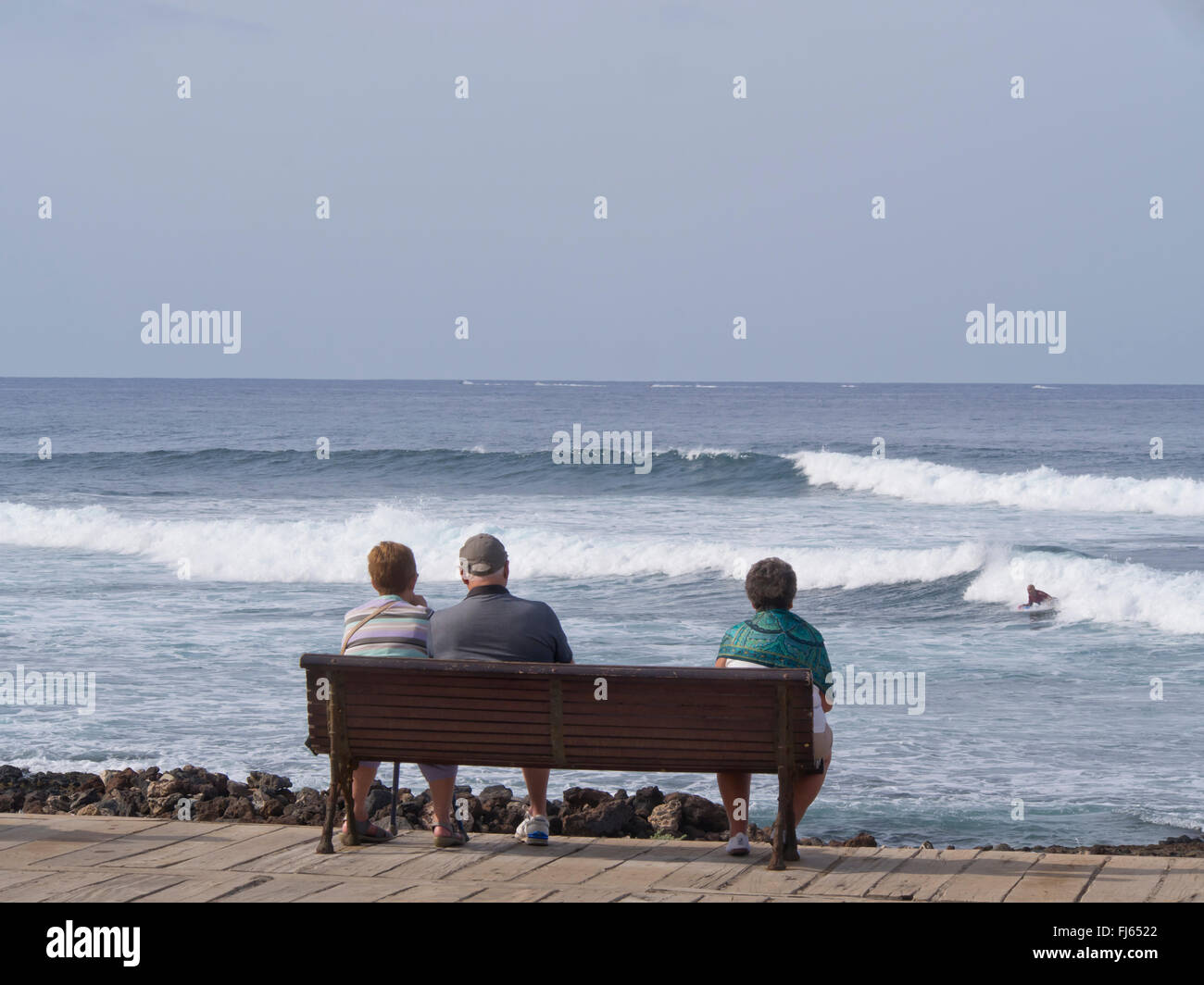 Tres ciudadanos de la tercera edad en un banco del paseo costero en Playa de las Americas Tenerife, mirando hacia el mar y las olas con surfer Foto de stock