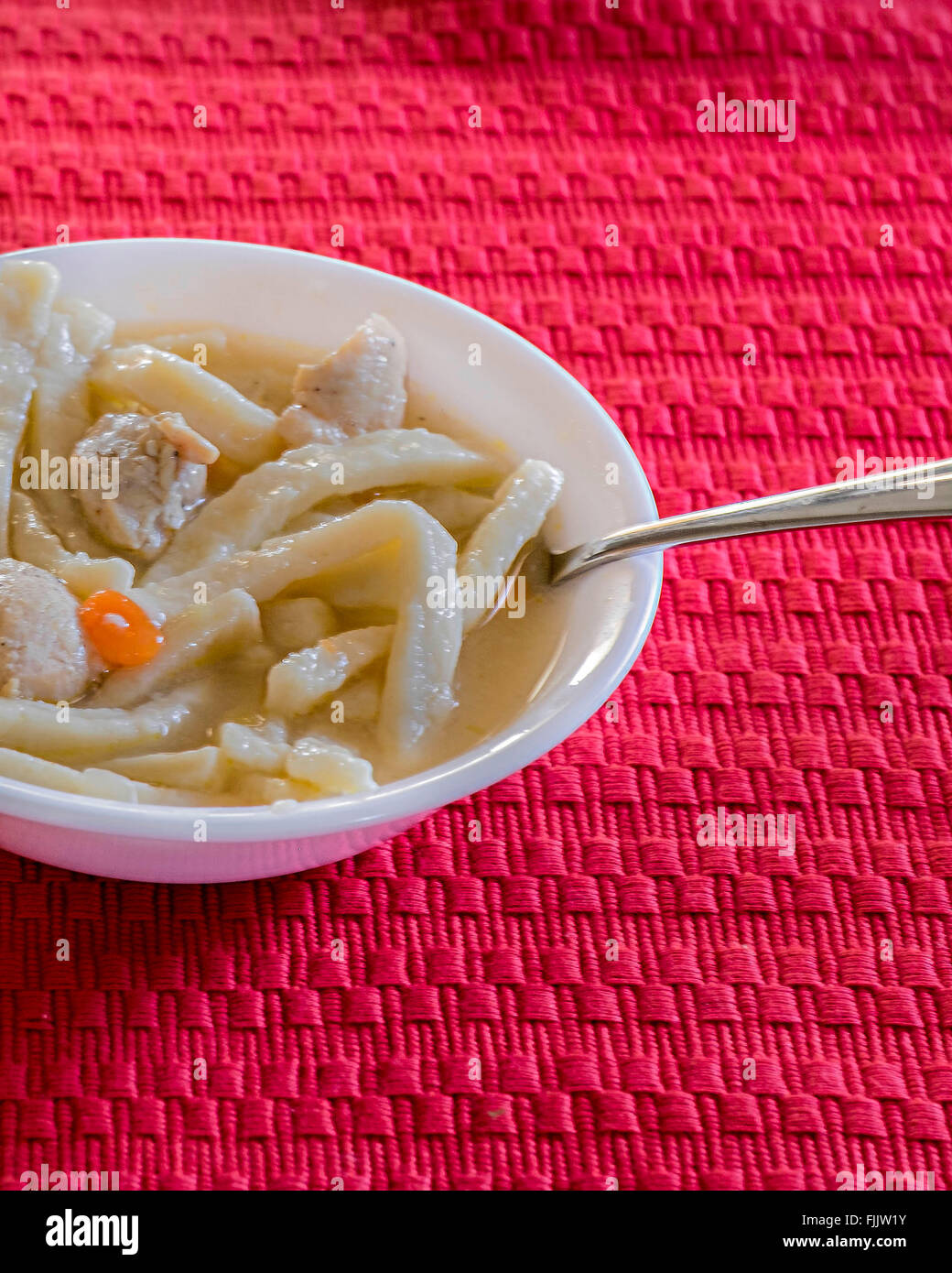 Un recipiente blanco lleno de pollo y fideos caseros sobre una alfombra roja. Foto de stock