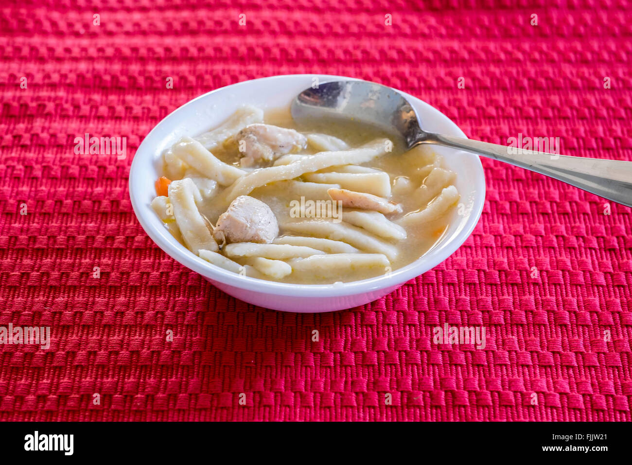 Un recipiente blanco lleno de pollo y fideos caseros sobre una alfombra roja. Oklahoma, Estados Unidos. Foto de stock