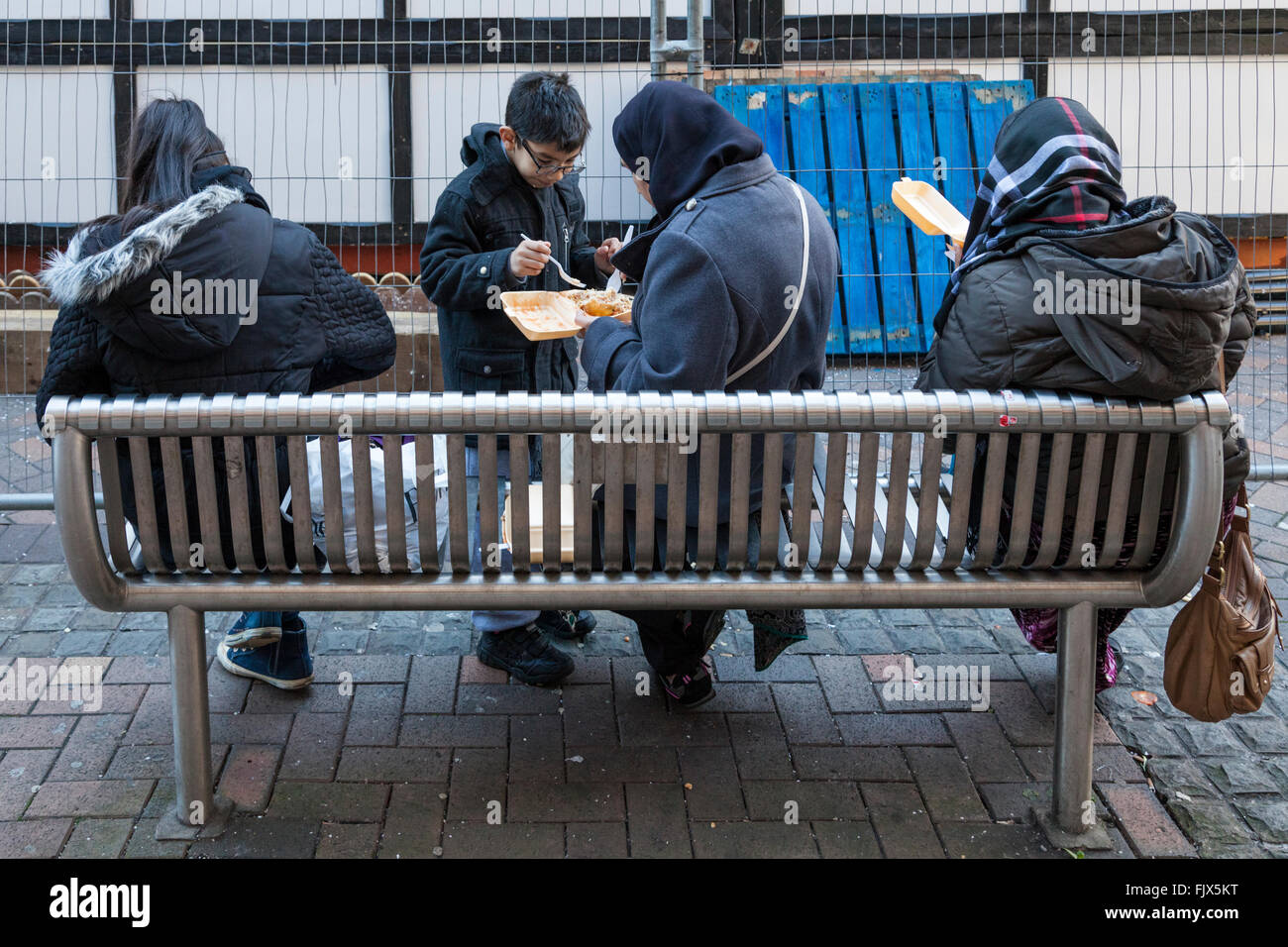 Familia sentada en un banco comiendo comida para llevar afuera en un día frío en el centro de la ciudad. Nottingham, Inglaterra, Reino Unido. Foto de stock