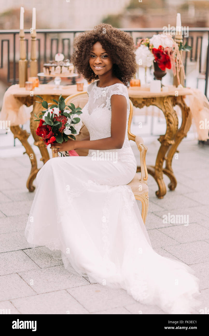 Feliz joven novia negra sonriente con bouquet de flores rojas y sentado en  la silla de terracota vintage Fotografía de stock - Alamy