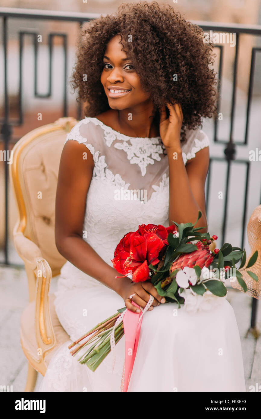 Feliz joven novia negra sonriente con bouquet de flores rojas y sentado en  la silla de terracota vintage Fotografía de stock - Alamy