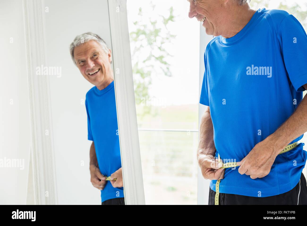 Feliz senior hombre midiendo su cintura mientras se mira en el espejo Foto de stock