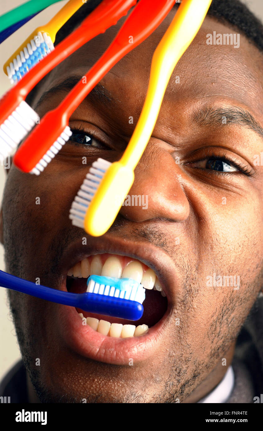 Un hombre joven pinceles sus dientes con una mano, mientras se mantiene una serie de cepillos de dientes en su otra mano. Foto de stock