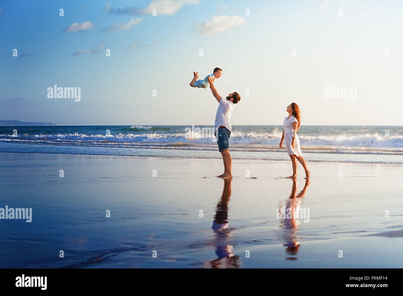 Familia Feliz, padre, madre, hijo camine con diversión a lo largo del borde del mar sunset surf en la playa de arena negra. Foto de stock