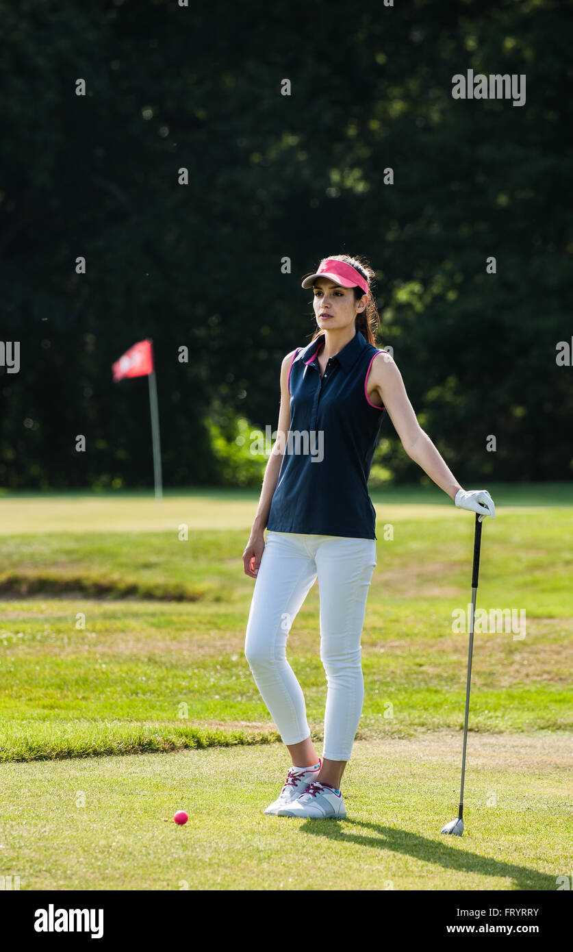 de hermosa mujer mirando el verde del de golf. Ella está esperando delante de su bola rosa, sosteniendo su club en un conjunto de ropa deportiva y una gorra