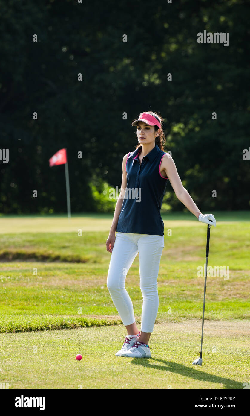 Retrato de una hermosa mujer mirando el verde del campo de golf. Ella está  esperando delante de su bola rosa, sosteniendo su club en un conjunto de  ropa deportiva y una gorra