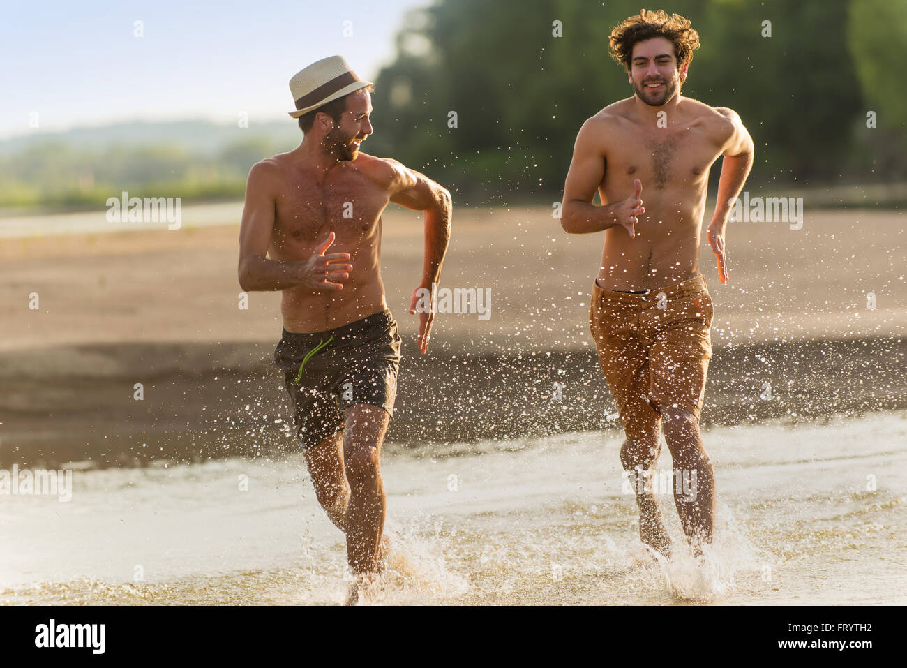 Dos amigos hombres se están ejecutando en el agua de la playa durante sus  vacaciones.Son descamisados, vistiendo trajes de baño y sombrero. El agua  salpica alrededor de ellos. Filmación con retroiluminación Fotografía