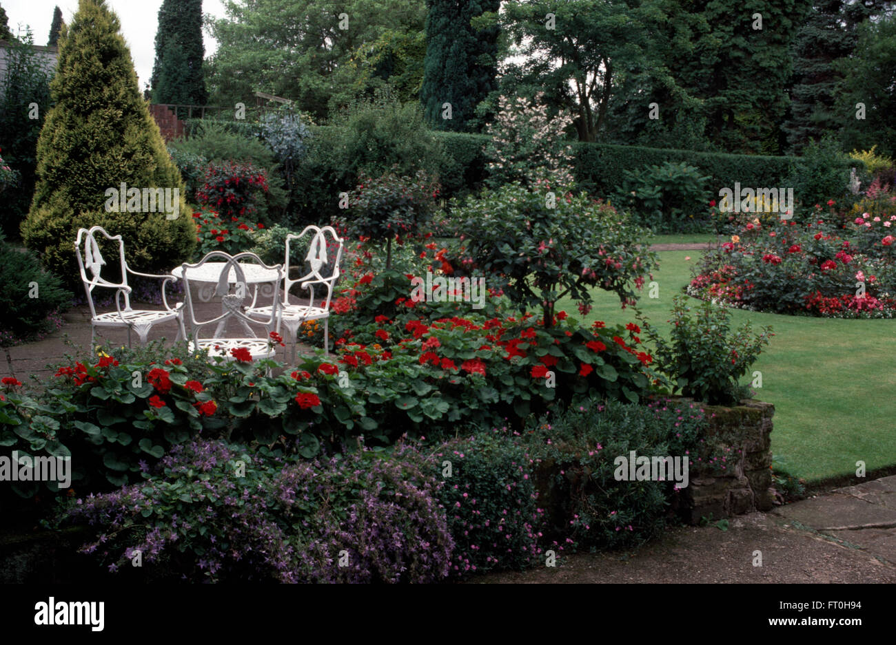 Blanco mesa y sillas de hierro forjado en el patio con un borde de color rojo fucsia y geranios en un jardín bien cuidados. Foto de stock