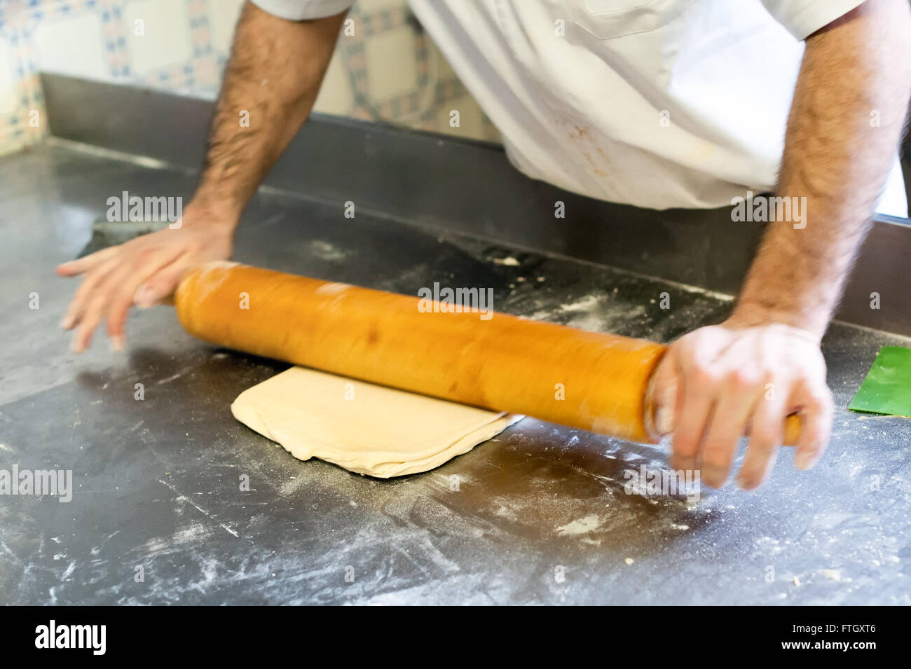 Cerrar la acción de disparo Baker macho rodando la masa con rodillo de madera sobre la superficie metálica en Configuración de panadería profesional Foto de stock