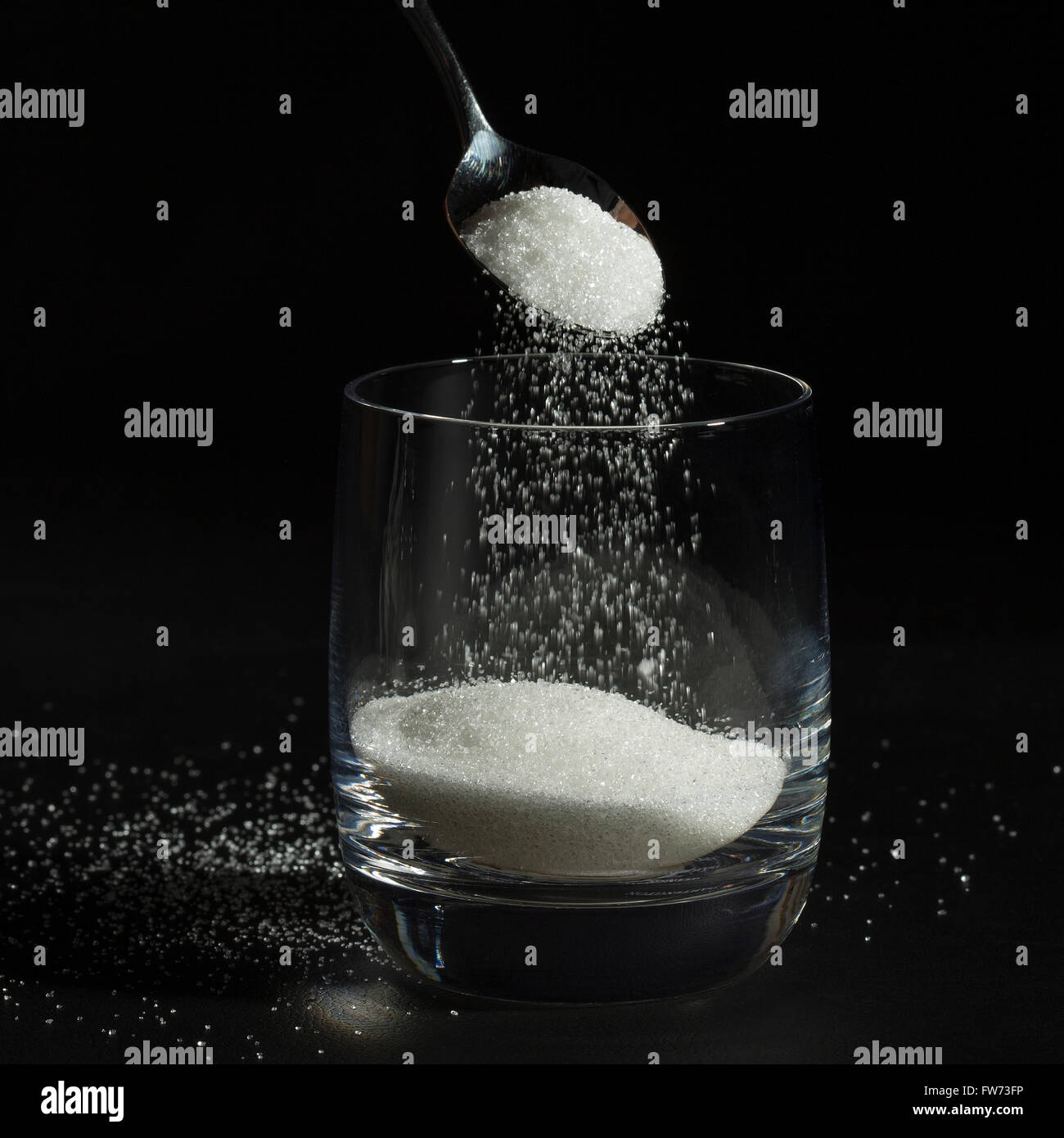 Caída de azúcar con una cuchara en un vaso en formato cuadrado, demostrando la cantidad excesiva de azúcar poner en muchas bebidas Foto de stock