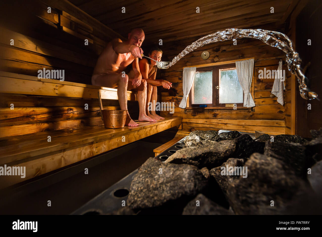 Pareja de mediana edad en madera tradicional sauna finlandesa, hombre echándole agua a la estufa caliente Foto de stock