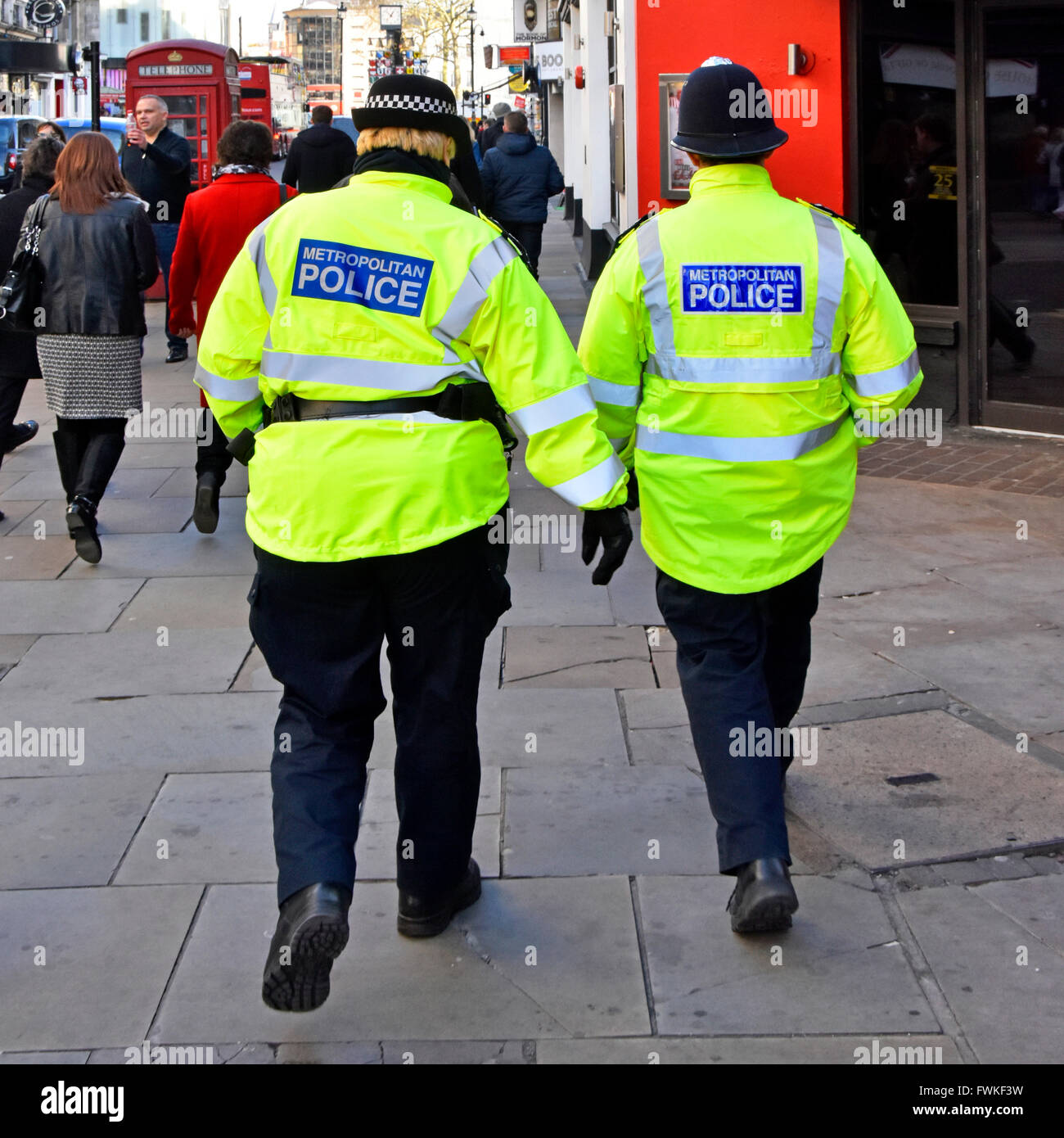 Vista posterior WPC funcionaria de la Policía Metropolitana en distintivo tapa (izquierda) y funcionario varón casco tradicional patrullaje West End de Londres Inglaterra Foto de stock