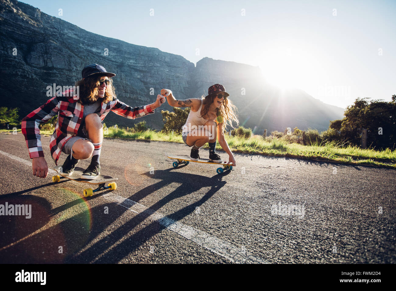 Feliz pareja joven divirtiéndose con el monopatín en la carretera. Hombre y mujer joven patinando juntos en un día soleado. Foto de stock