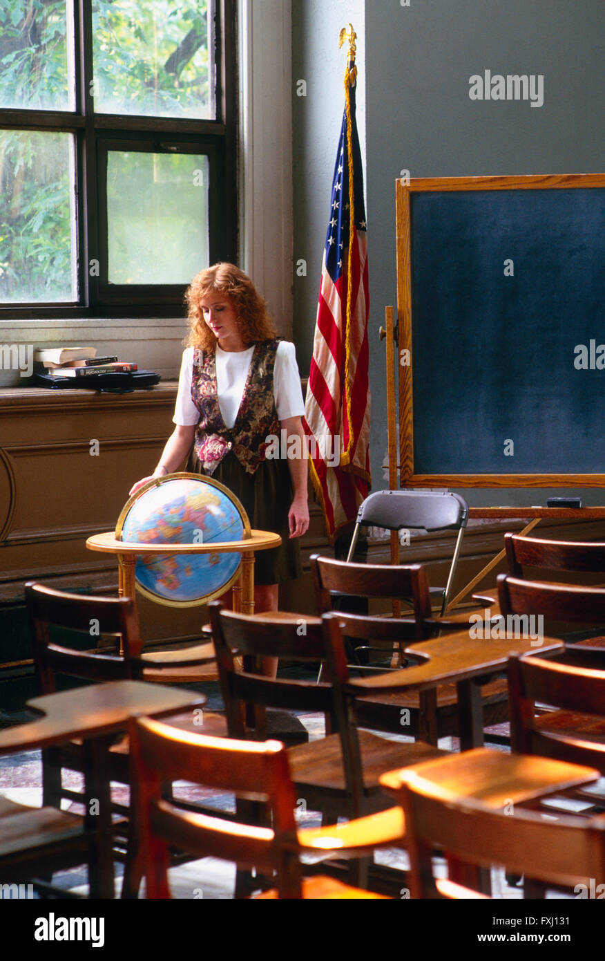 Red Headed femenina docente en aula con globo terráqueo, Bandera Americana y pizarra Foto de stock