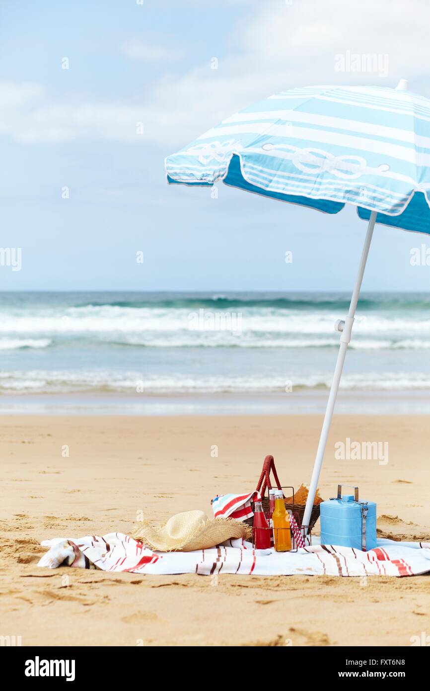 Sombrero, cool box y cesta de picnic en una toalla de playa debajo de una sombrilla en la playa Foto de stock