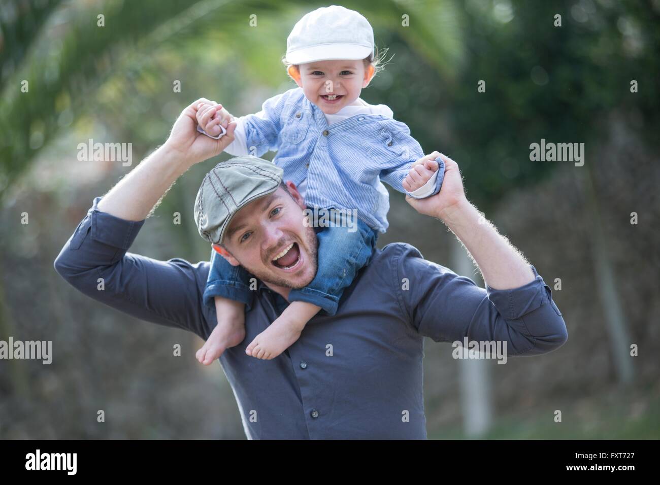 Baby Boy vistiendo una gorra de béisbol sentado en padre hombros mirando a la cámara sonriendo Foto de stock