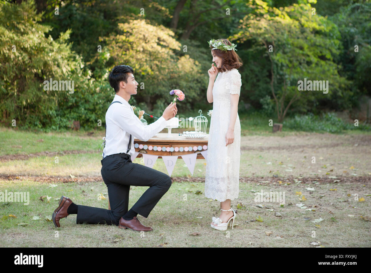 Esposo de rodillas y dando un ramo para la novia delante de un juego de mesa para boda fuera Foto de stock
