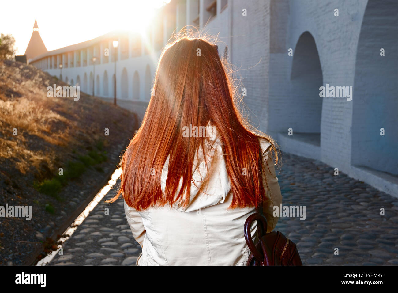 Vista trasera de la mujer de pelo rojo caminando por la calle cerca de la vieja fortaleza de Europa retroiluminados con sunflares Foto de stock