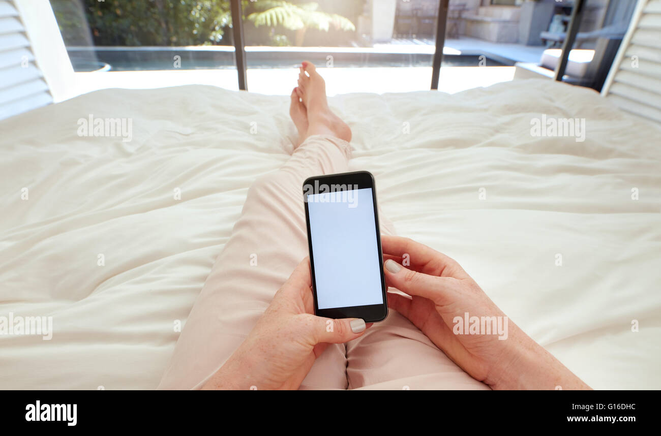 Closeup imagen de mujer acostada en una cama sosteniendo un teléfono inteligente con pantalla en blanco. POV shot de mujer relajante en la habitación utilizando la pantalla táctil Foto de stock