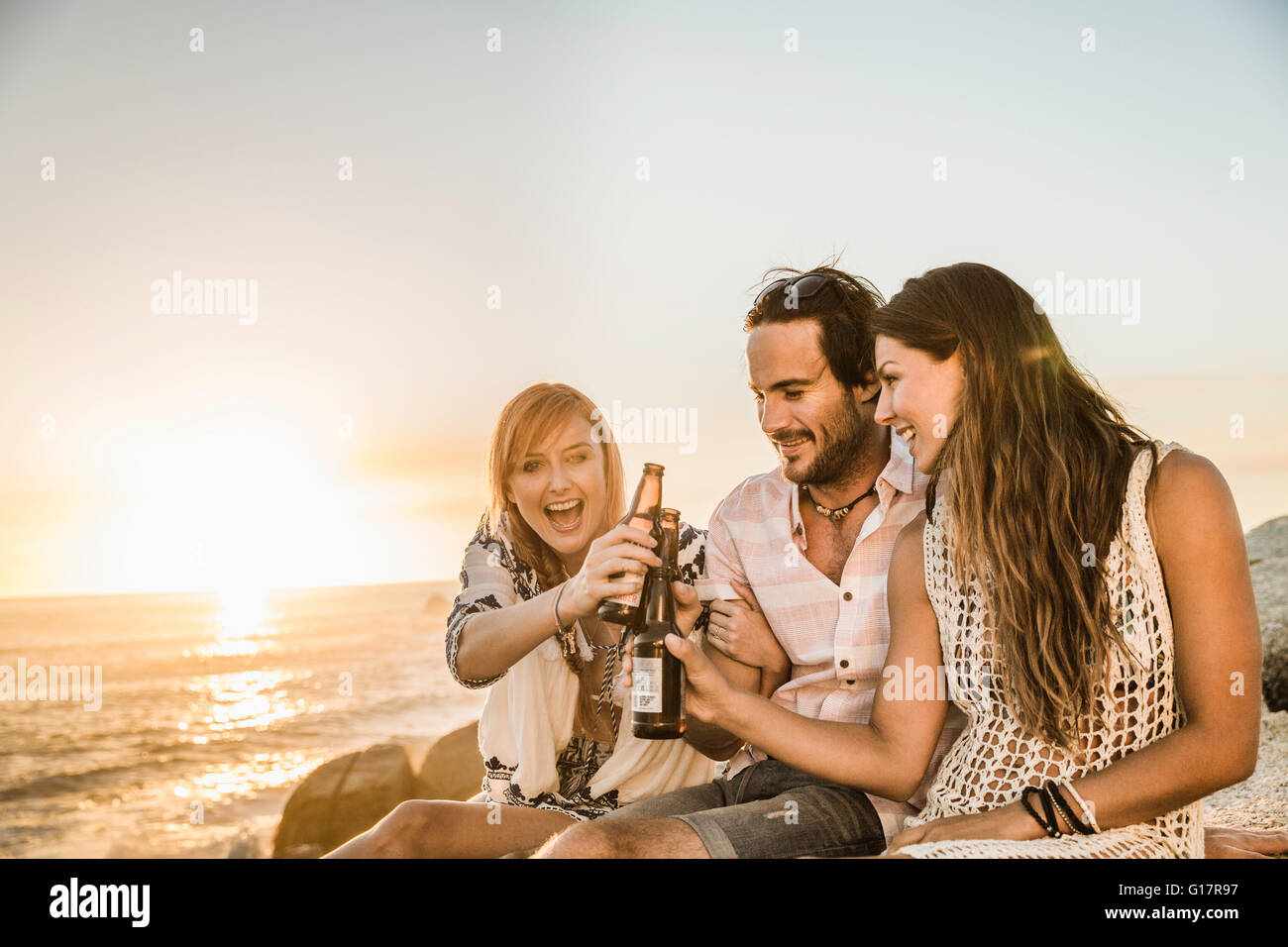 Tres mediados de amigos adultos sentados en la playa al atardecer, hacer un brindis, Ciudad del Cabo, Sudáfrica Foto de stock