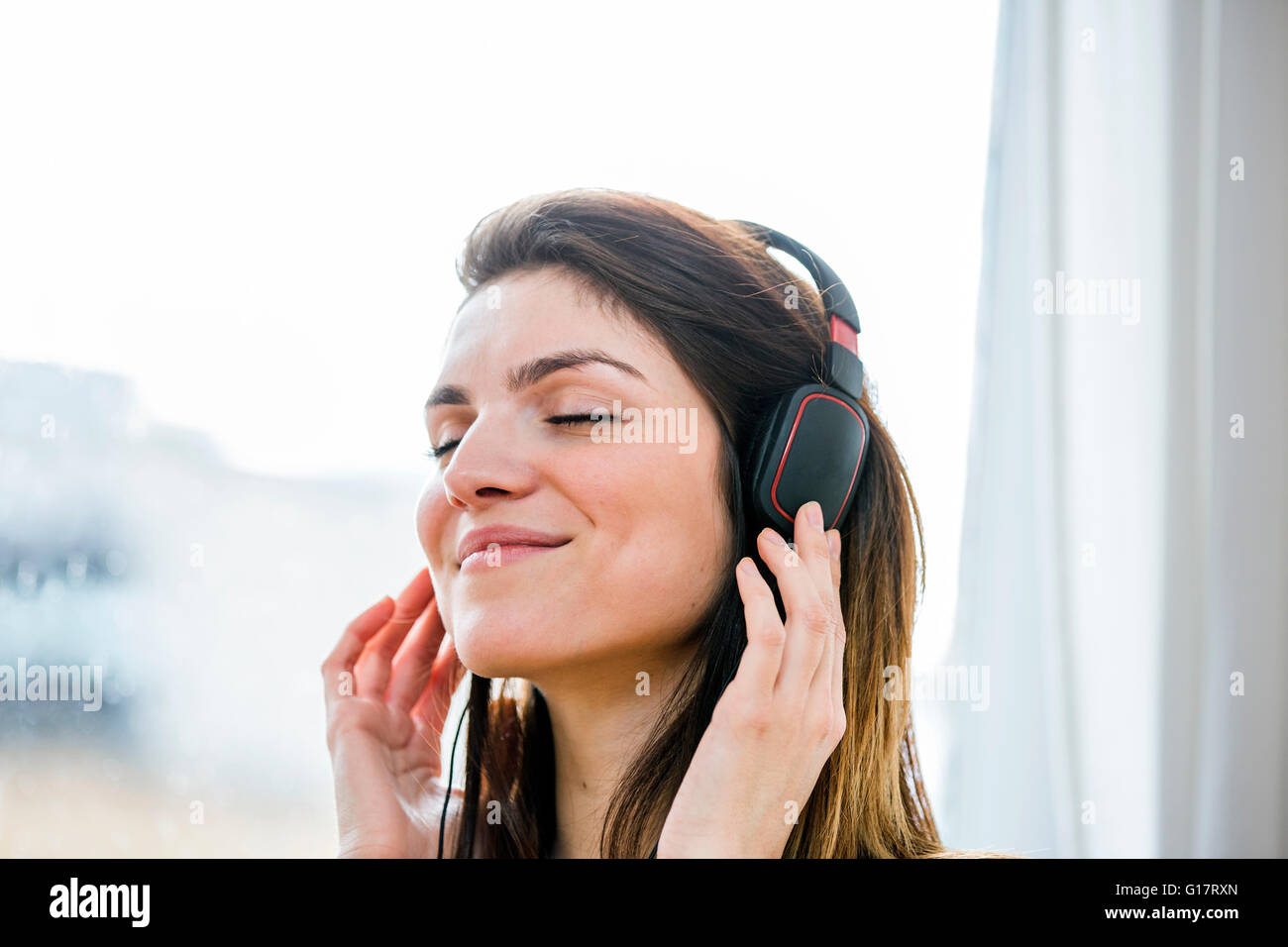 Hermosa joven escuchando música con auriculares en frente del apartamento de la ventana Foto de stock