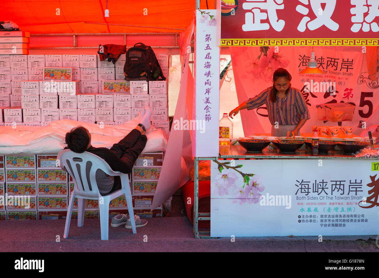 Hombre chino a dormir en el lugar de trabajo en un mercado, mientras que su vecino femenino está trabajando Foto de stock