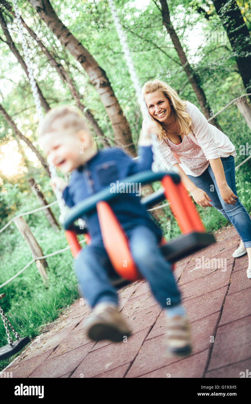 Madre e hijo al aire libre en el patio de recreo cabalgando en un columpio y sonriente Foto de stock