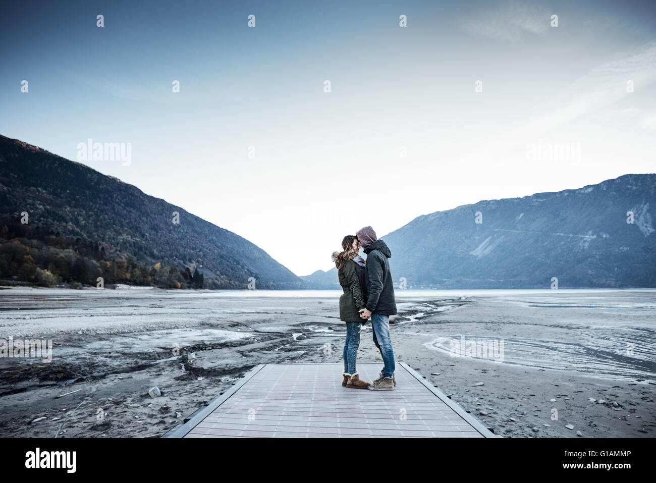 Joven pareja romántica en un muelle cogidos de la mano, el lago y las montañas al fondo Foto de stock