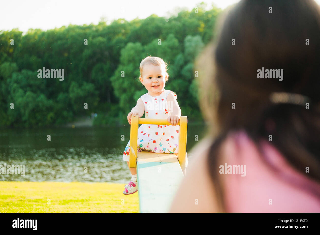 Niña disfruta montando swing con mamá por la bella naturaleza de fondo. Bebé feliz jugando con los padres fuera Foto de stock