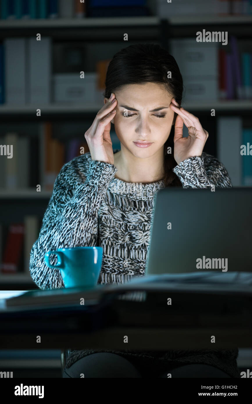Cansado joven utilizando un ordenador portátil y conexión a internet a altas horas de la noche, ella está teniendo un dolor de cabeza y tocando su compra Foto de stock