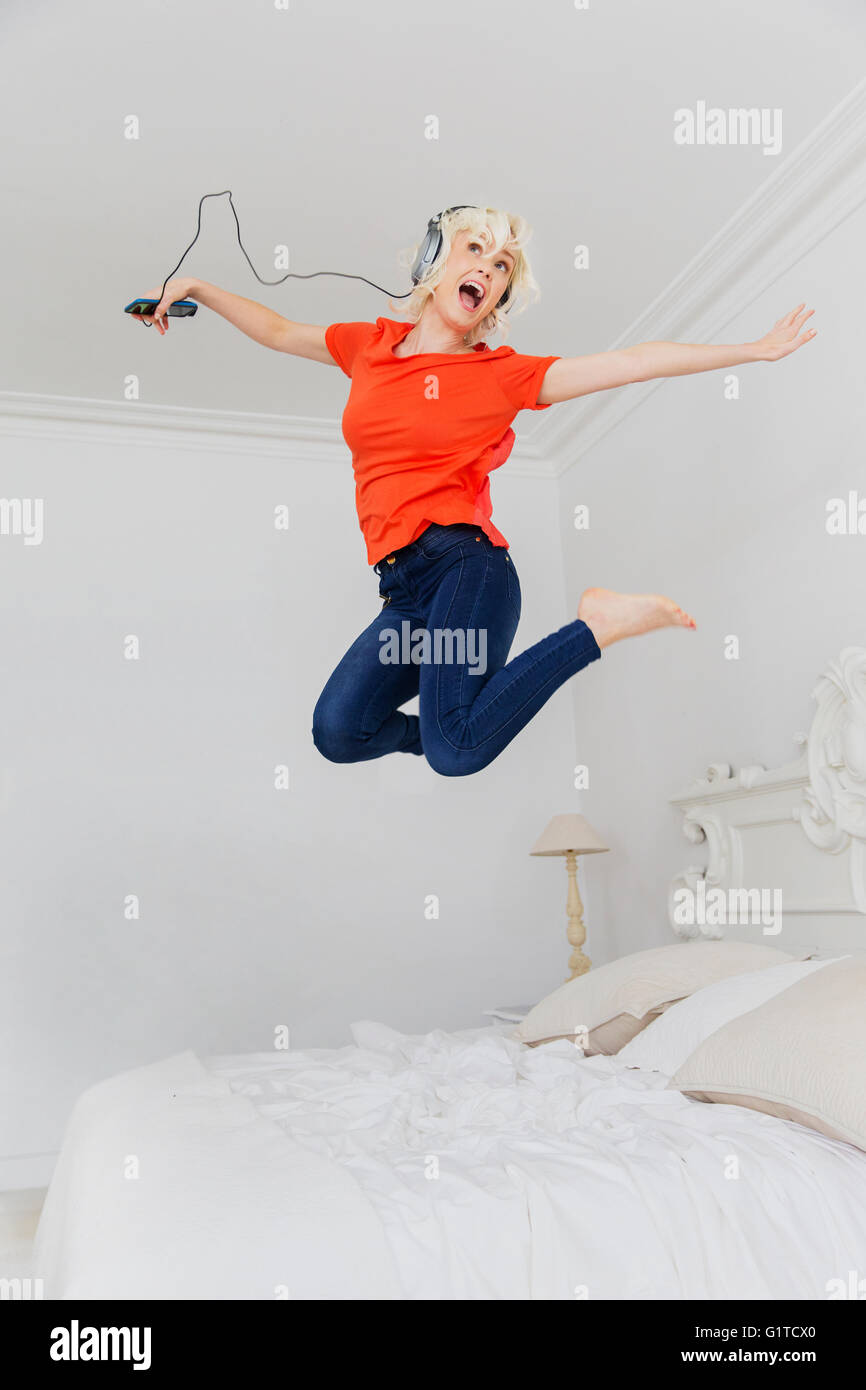 Mujer juguetona saltando sobre la cama escuchando música con auriculares y reproductor mp3 Foto de stock