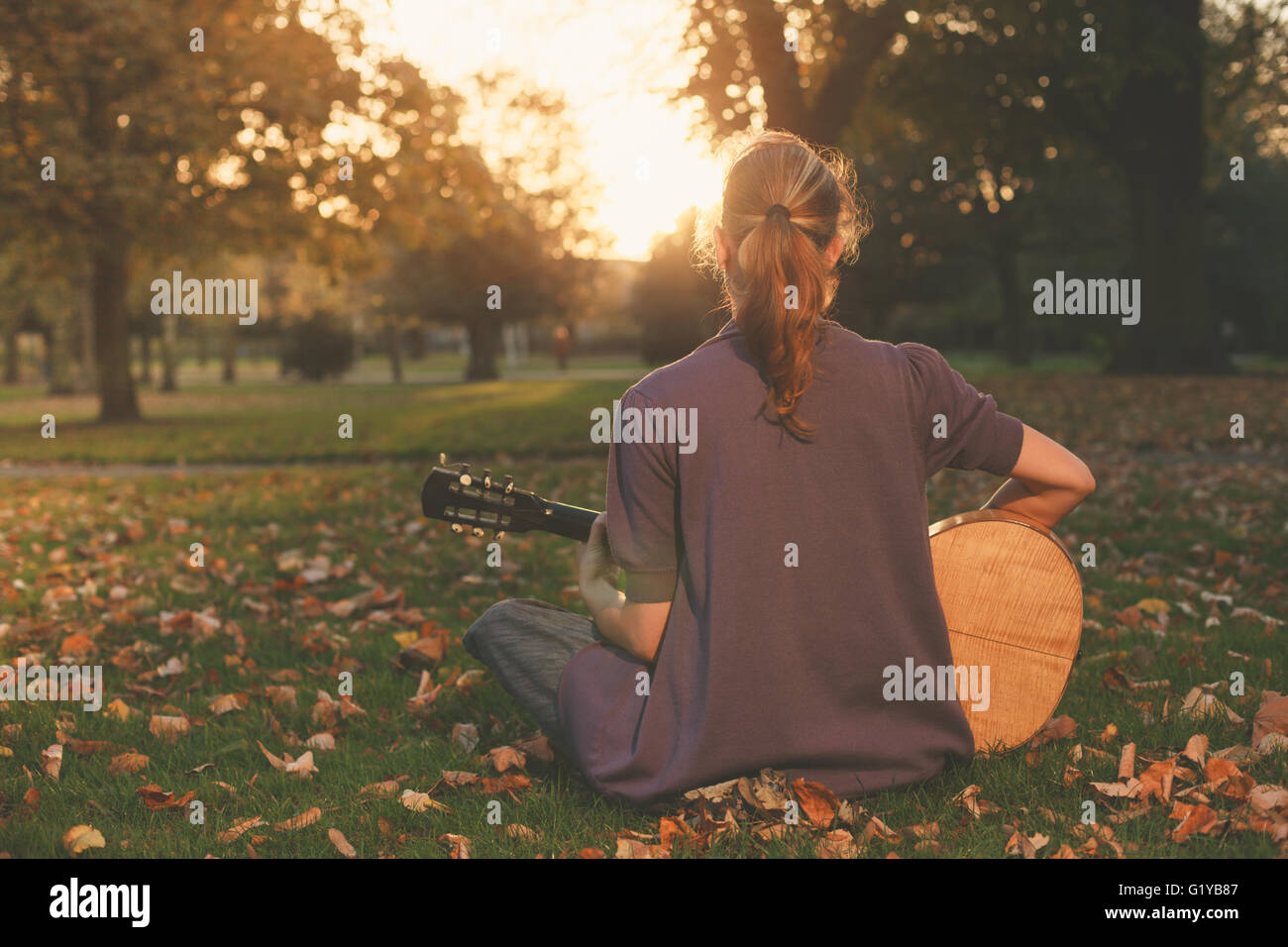 Vista trasera de una joven sentada sobre el césped y tocando la guitarra en el parque al atardecer Foto de stock