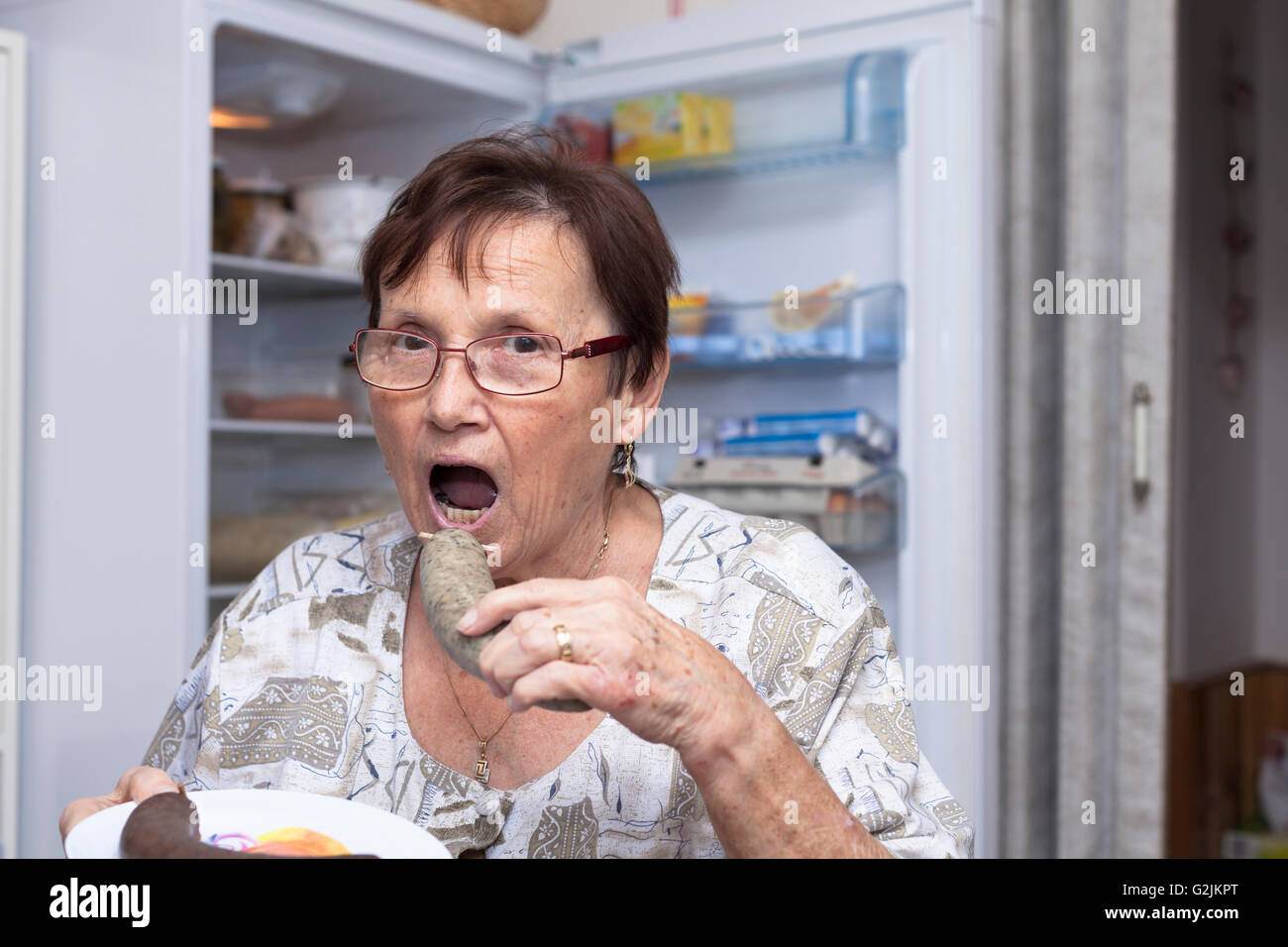 Mujer mayor va a comer carne de cerdo salchicha de hígado mientras está de pie delante de la nevera en la cocina abierta. Foto de stock