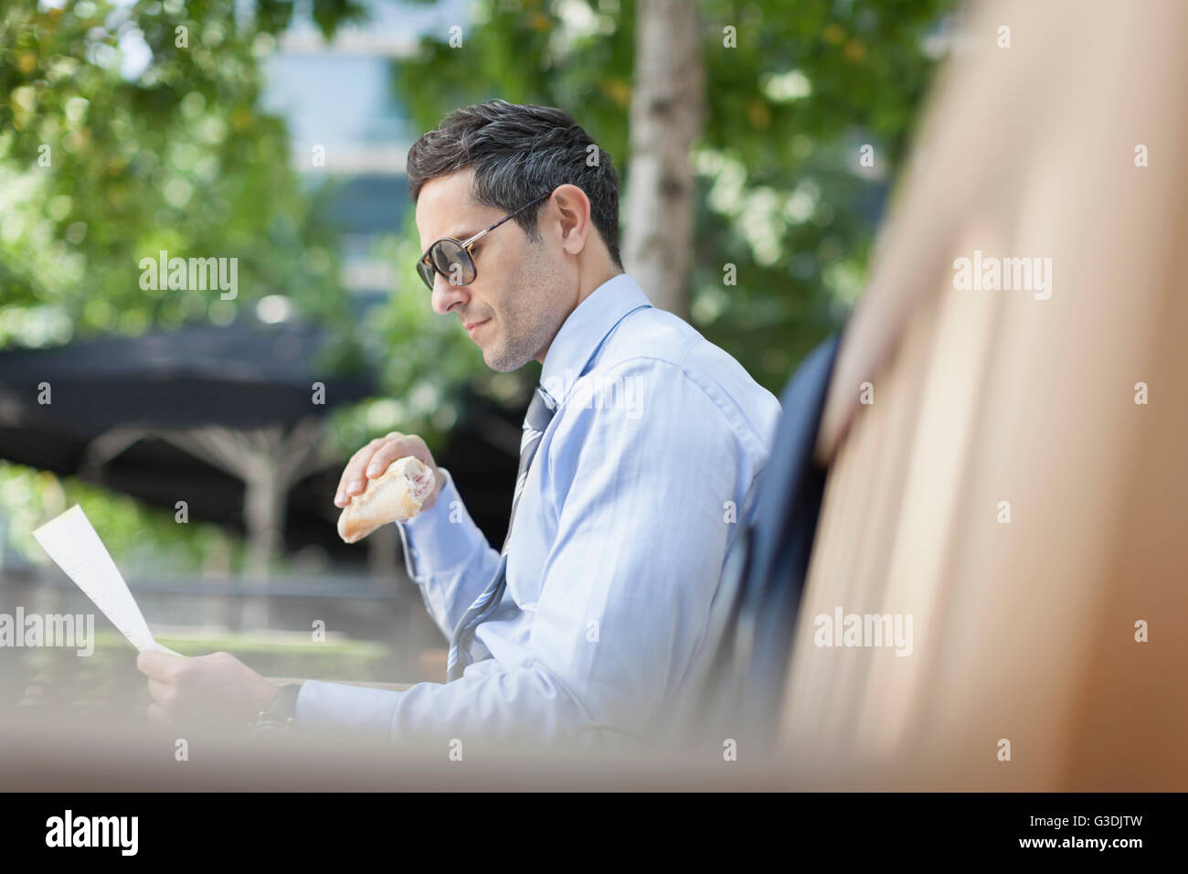 Empresario corporativo comiendo y leyendo el papeleo en un banco del parque Foto de stock