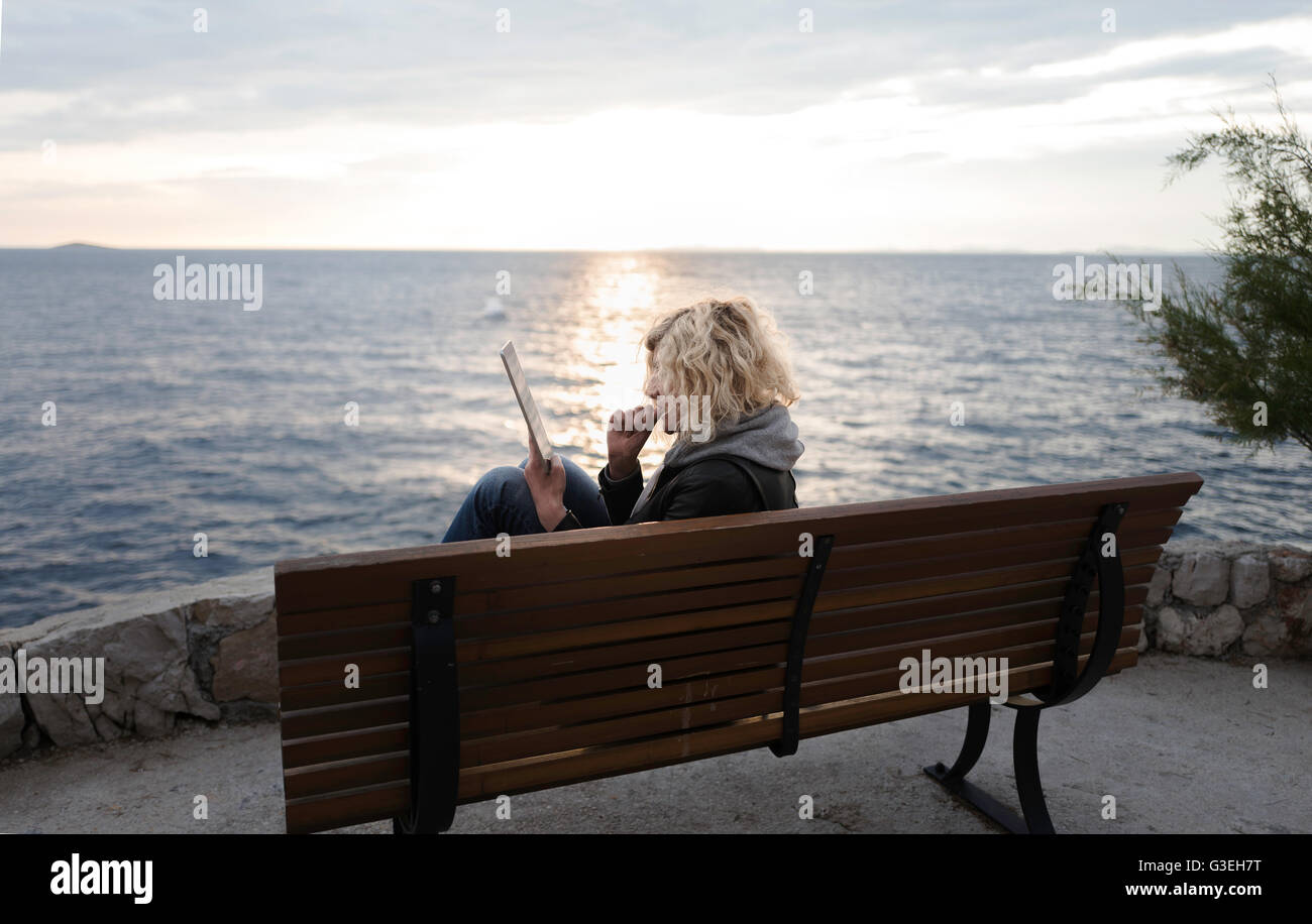 Chica rubia con el pelo rizado es sentarse en el banquillo y absorbedly viendo algo en tablet Foto de stock