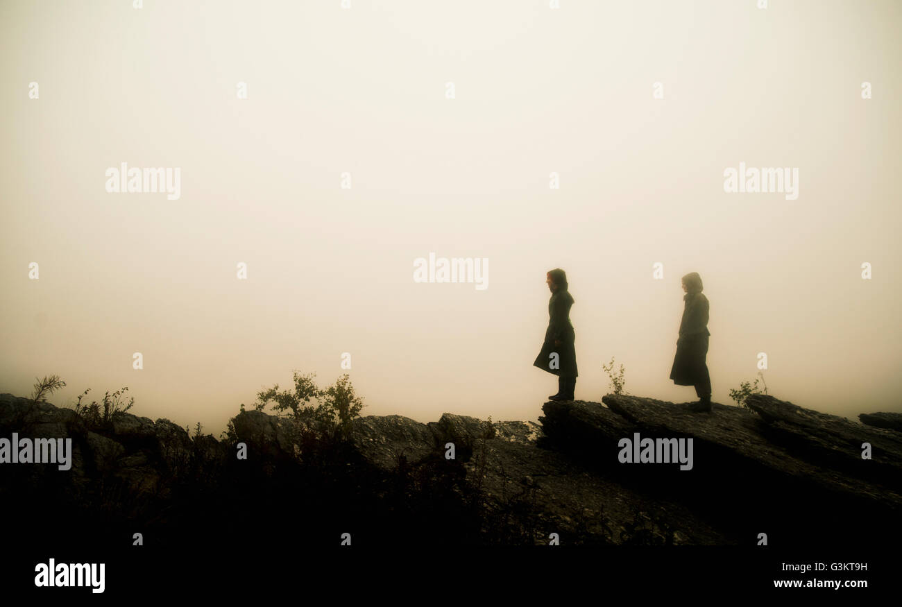 Vista lateral de la pareja vistiendo impermeables de pie sobre las rocas en la niebla en busca de distancia Foto de stock