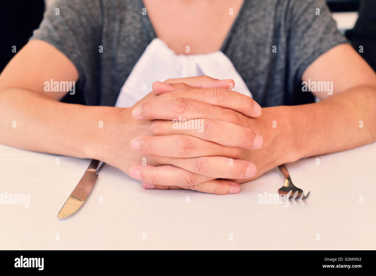 Primer plano de un joven hombre caucásico sentados en la mesa esperando la comida, con las manos entrelazadas y un cuchillo y un tenedor en la fro Foto de stock