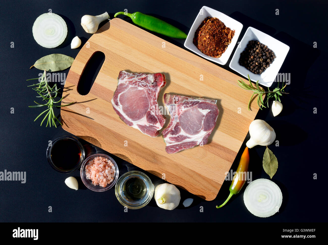 La carne de cerdo cruda sobre una tabla para cortar y pimienta, laurel, romero, cebolla, sal, aceite de oliva, salsa sobre un fondo negro Foto de stock