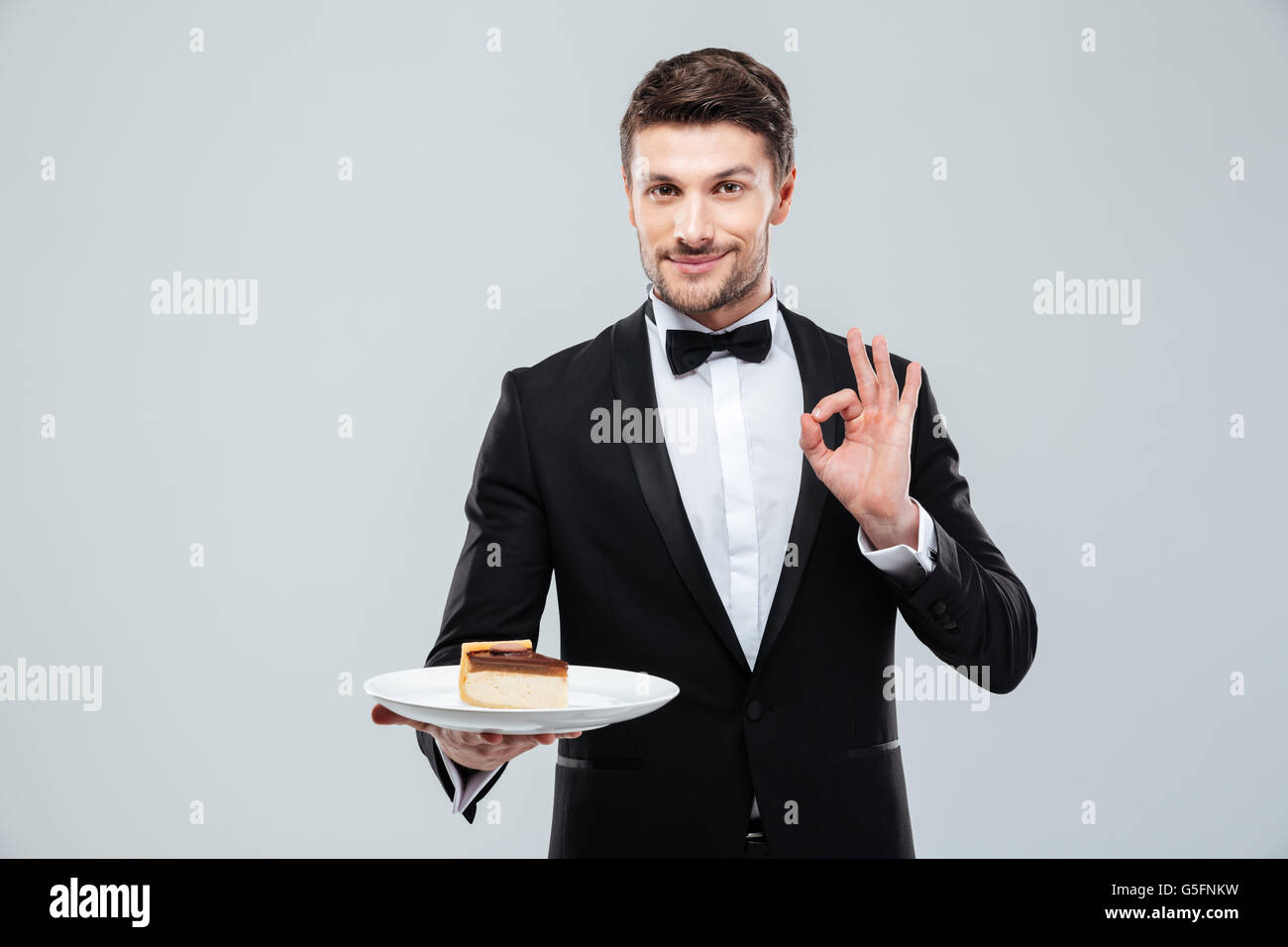 Apuesto joven camarero en smoking con pajarita que sujetan la placa con tarta y mostrando signo ok Foto de stock