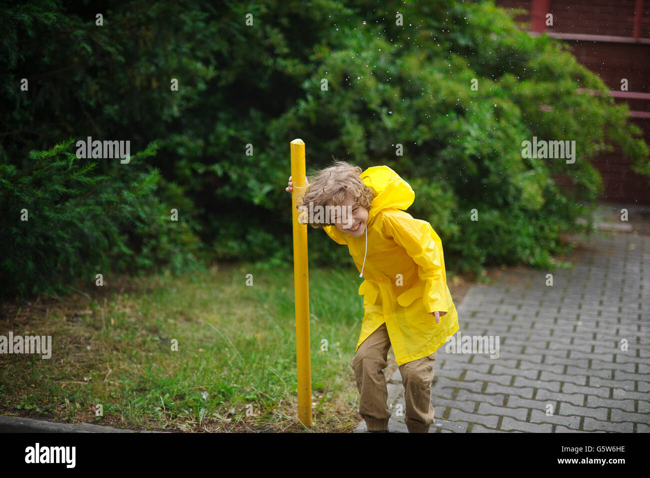 El chico en un impermeable amarillo está en la lluvia en el patio y se ríe fuerte. Frías gotas de lluvia sobre la cabeza. El muchacho