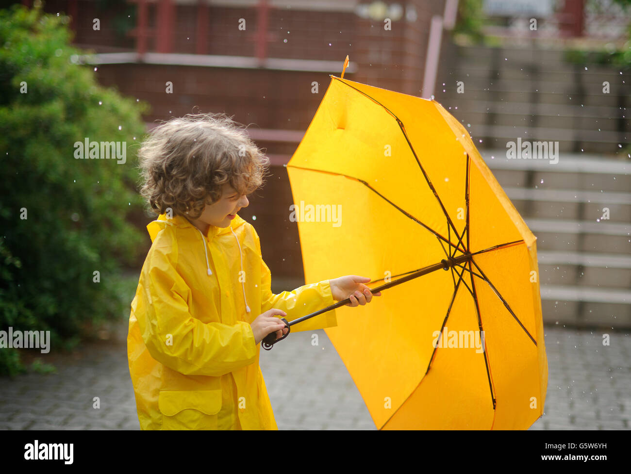El chico en un impermeable amarillo brillante abre un paraguas durante una lluvia. El niño está en el patio de la casa. Gotas de lluvia
