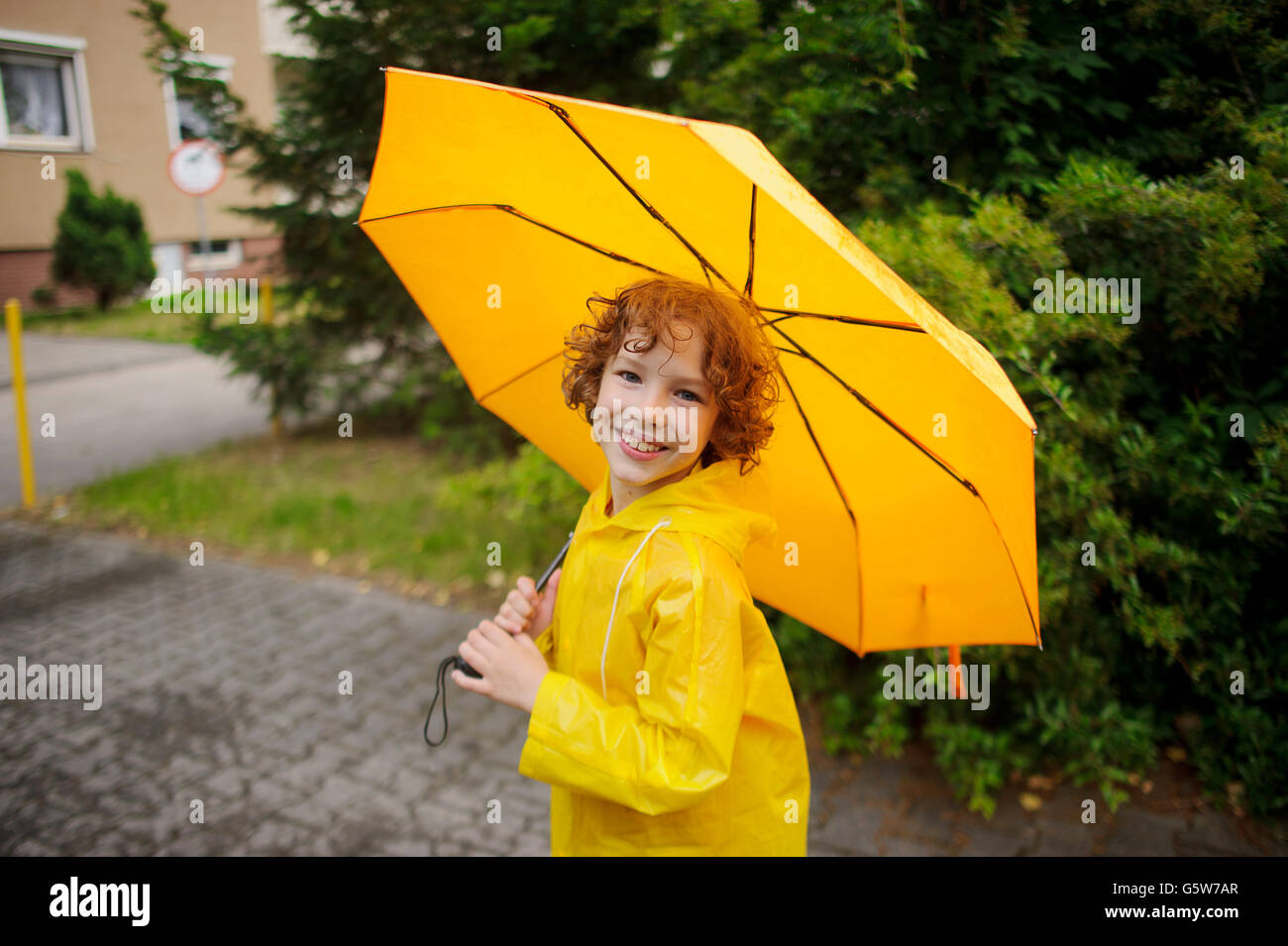 Alegre niño de 8-9 años bajo un paraguas amarillo. niño con una sonrisa busca en la cámara. Tiene el pelo rizado y húmedo una NIC Fotografía de stock -