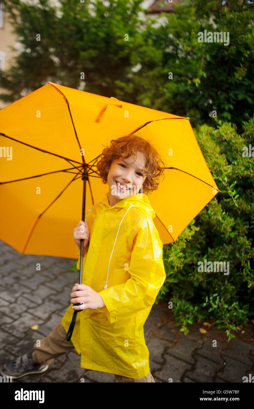 El chico lindo de 8-9 años bajo un paraguas amarillo. El chico en un impermeable amarillo brillante posee un gran paraguas en mano. El niño l