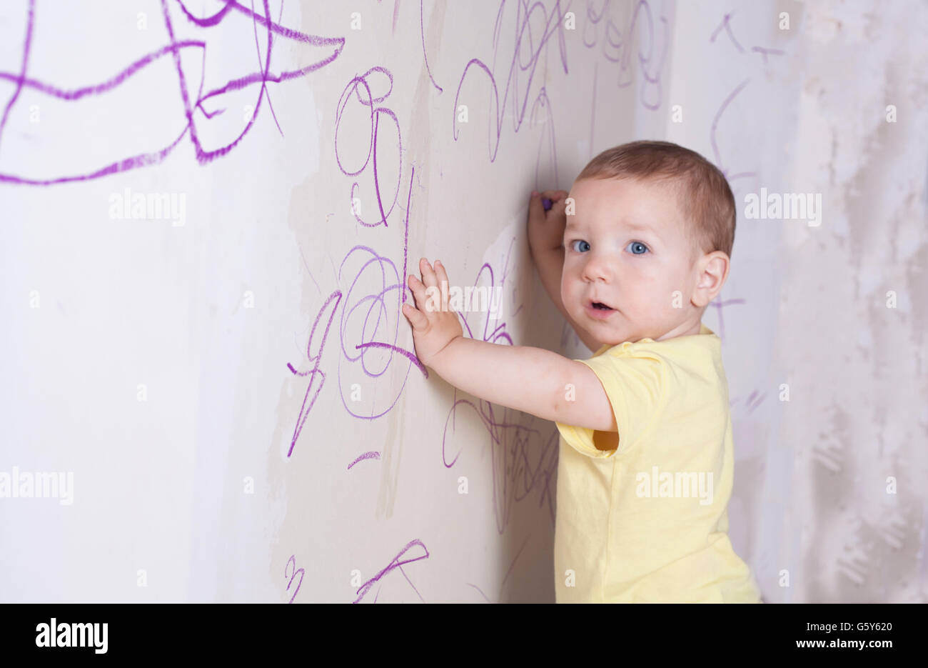 Baby Boy dibujo con lápiz de cera sobre la pared de yeso. Él está mirando a la cámara Foto de stock